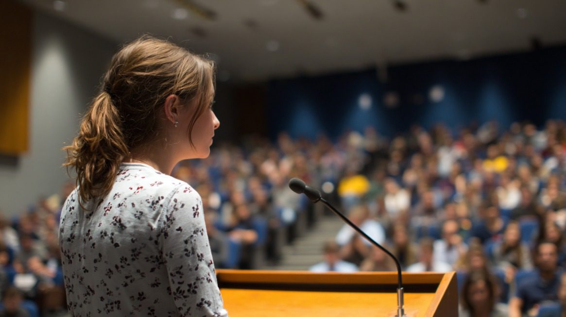 Young woman delivering a speech at a podium in front of a large audience