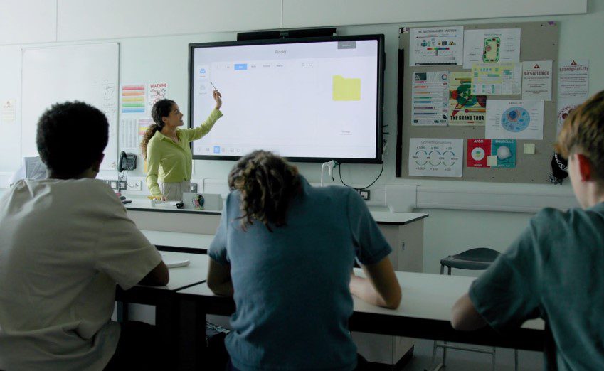 A teacher instructs a diverse group of students seated at desks in a bright classroom