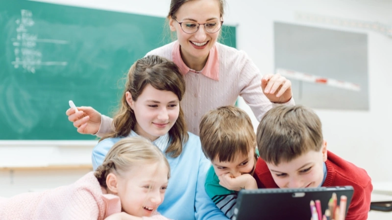 A teacher and students smile as they look at a tablet together during a classroom activity