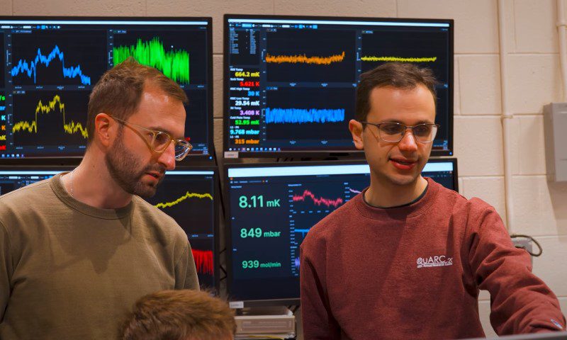 Two scientists intently observe multicolored data graphs displayed across multiple monitors in a laboratory setting