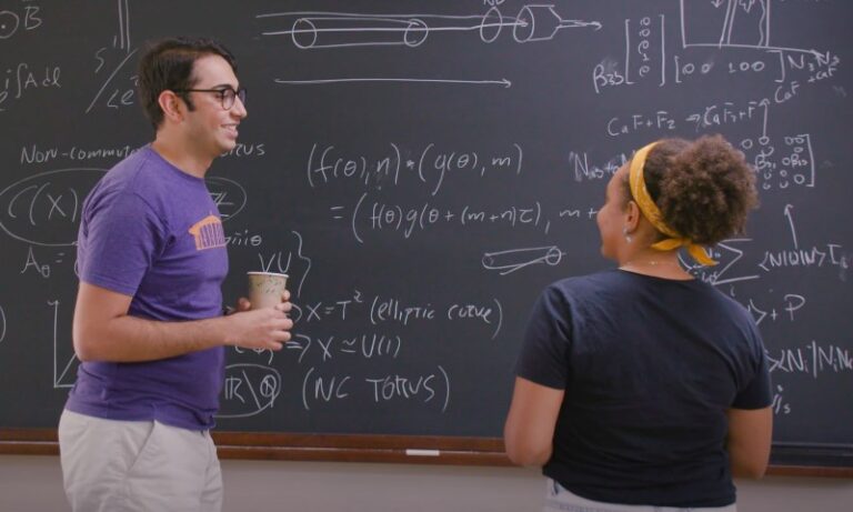 Two individuals discussing complex mathematical formulas written in white chalk on a dark blackboard