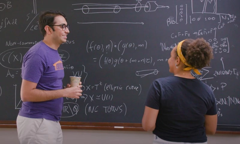 Two individuals discussing complex mathematical formulas written in white chalk on a dark blackboard
