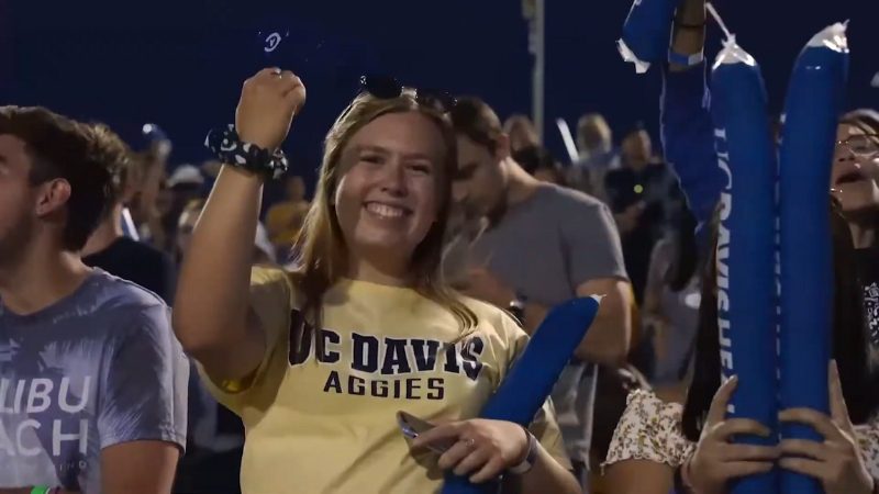 A UC Davis student cheers during a campus event, wearing a UC Davis Aggies shirt and holding blue foam sticks at the stadium